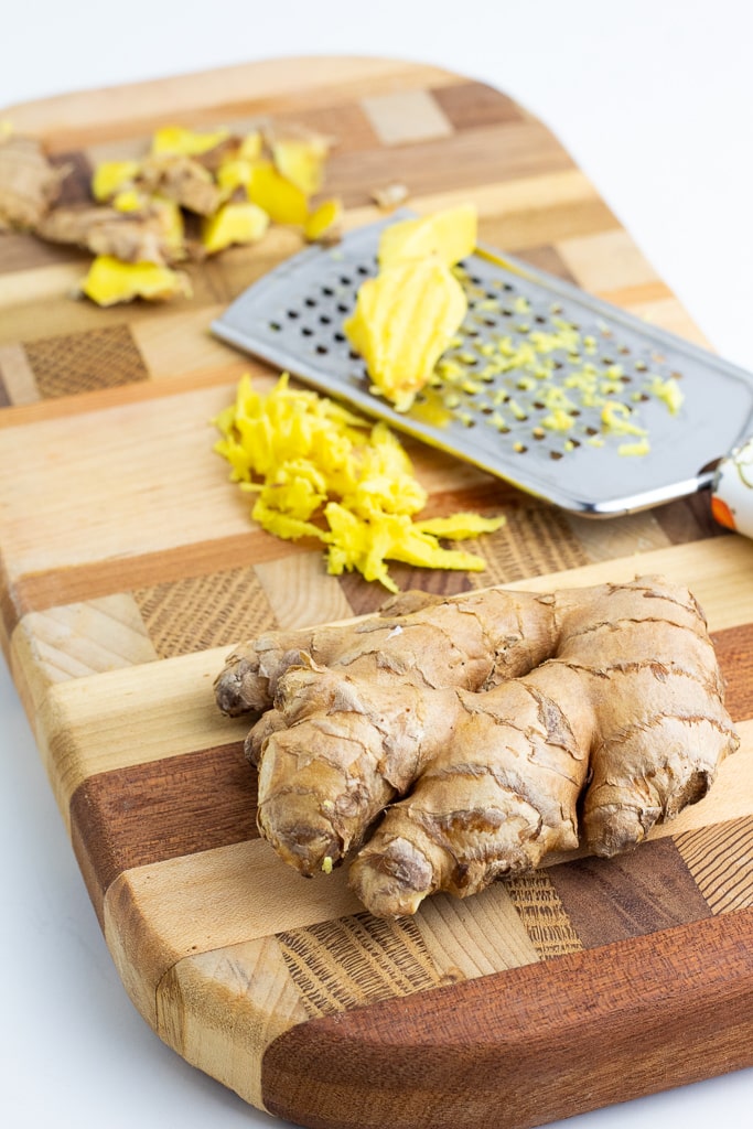 ginger root on cutting board with piece being grated in background