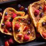 vegan french toast on a black platter and topped with strawberries, blueberries, and maple syrup.