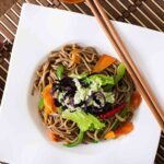 soba noodle veggie stir fry on a white square plate with wooden chopsticks.