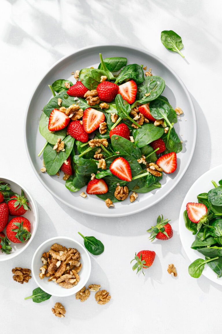 white plate of spinach salad with strawberries and walnuts on white table