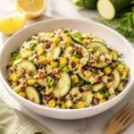 white bowl filled with zuchini corn salad with sliced lemon and zuchinni in background.