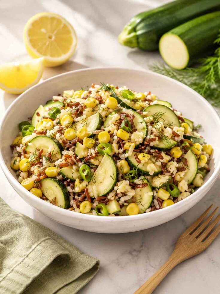 white bowl filled with zuchini corn salad with sliced lemon and zuchinni in background.