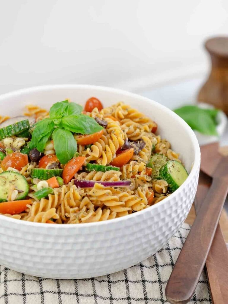 vegetable pasta salad in a white textured bowl topped with basil leaves.