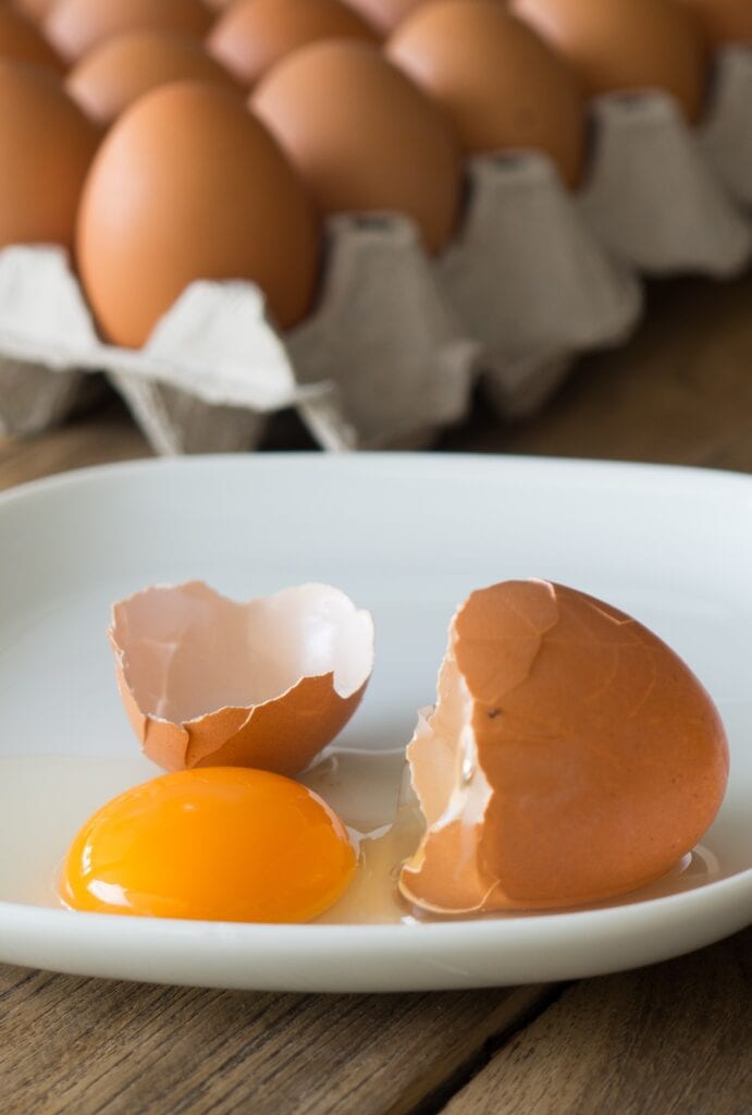cracked brown egg on white plate with crate of eggs behind