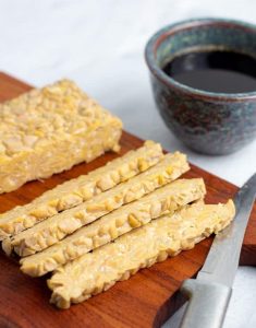 Sliced tempeh on wooden board with cup of marinade in background