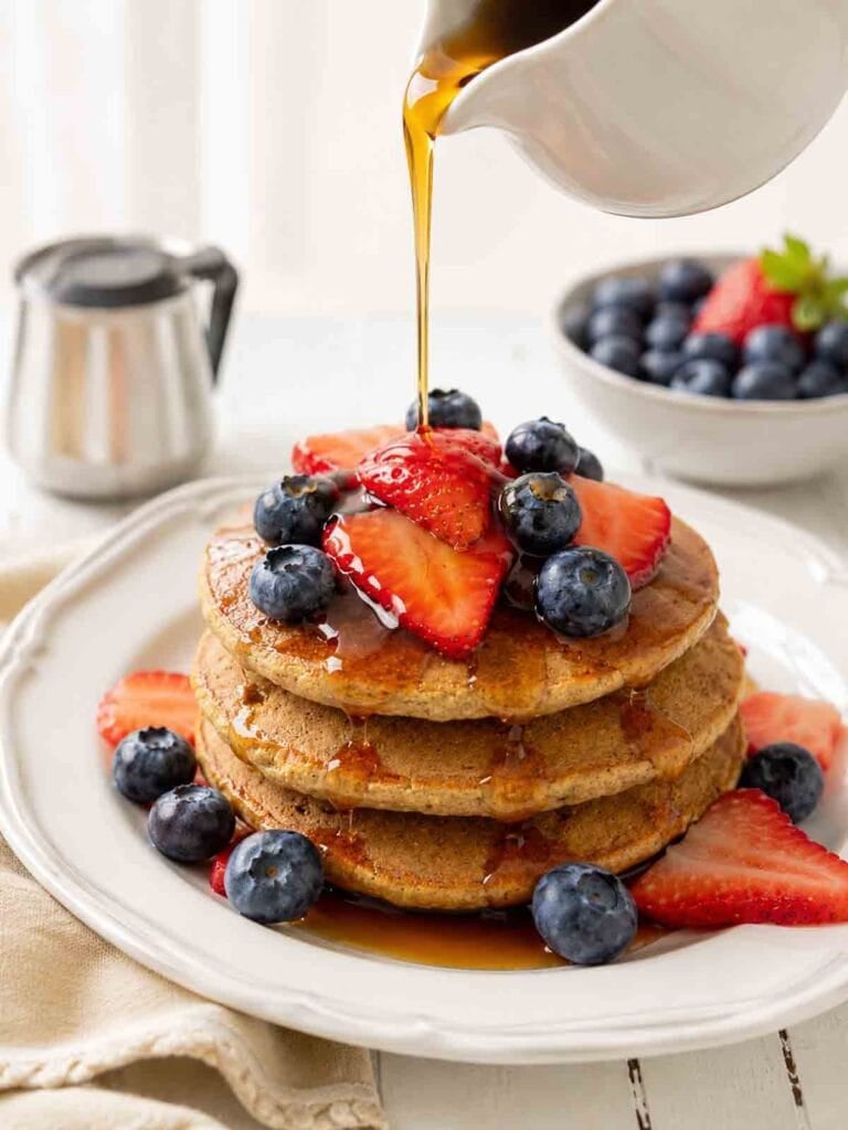 a stack of homemade pancakes topped with fruit and maple syrup being poured over top.