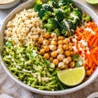 colorful veggie bowl with brown rice, chickpeas, steamed broccoli, shredded broccoli, shredded carrots, and tahini sauce.