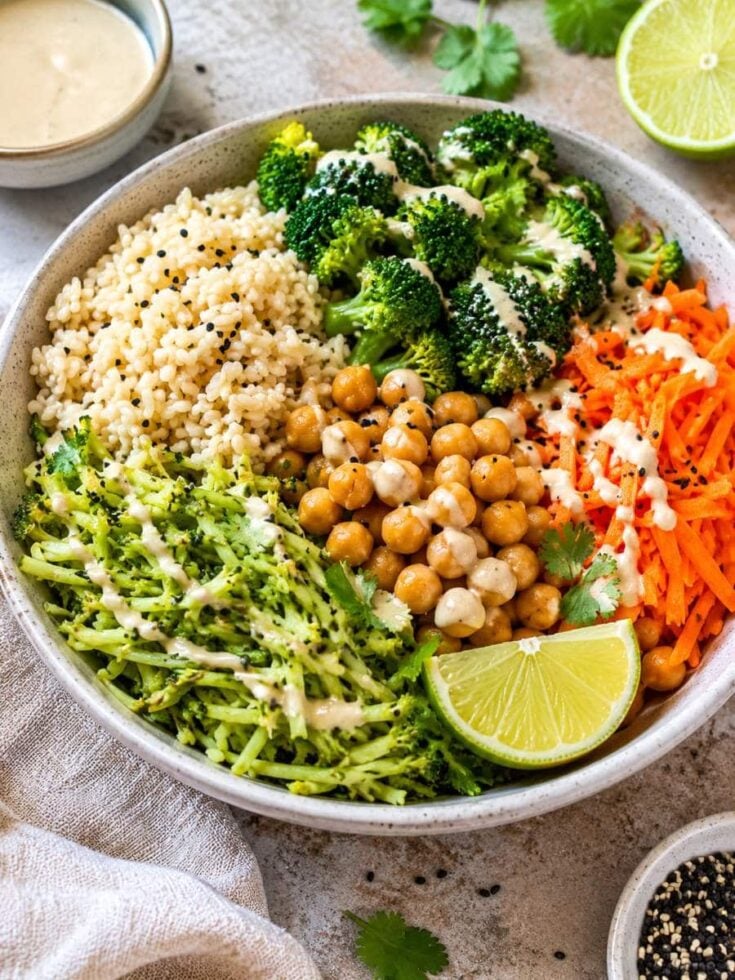 colorful veggie bowl with brown rice, chickpeas, steamed broccoli, shredded broccoli, shredded carrots, and tahini sauce.