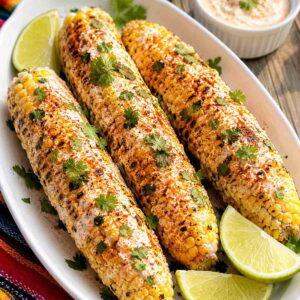 mexican street corn on the cob on a white platter with lime wedges.
