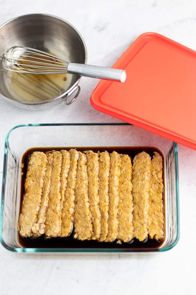 Sliced tempeh in glass dish marinating on white background
