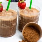 two vegan chocolate banana smoothies in glasses with green straws and strawberries, cocoa powder spilled on white background