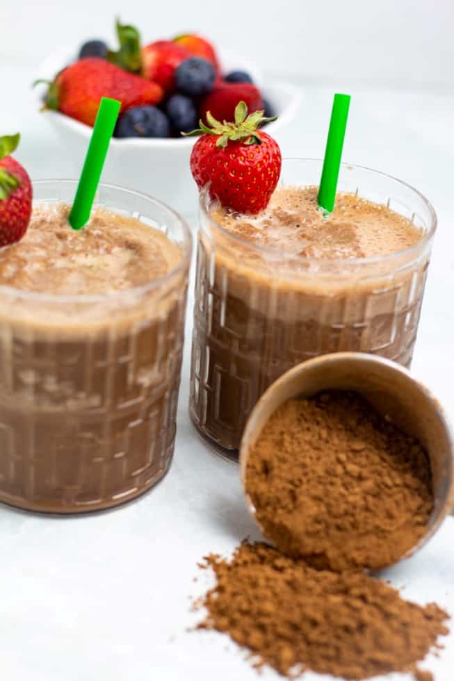 two vegan chocolate banana smoothies in glasses with green straws and strawberries, cocoa powder spilled on white background