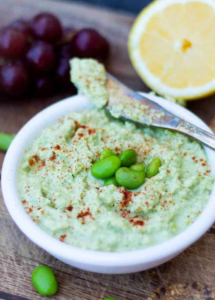 edamame hummus topped with whole bright green edamame beans in white bowl with spreading knife