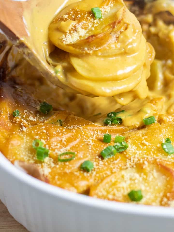 close up of vegan scalloped potatoes on wooden spoon over casserole dish