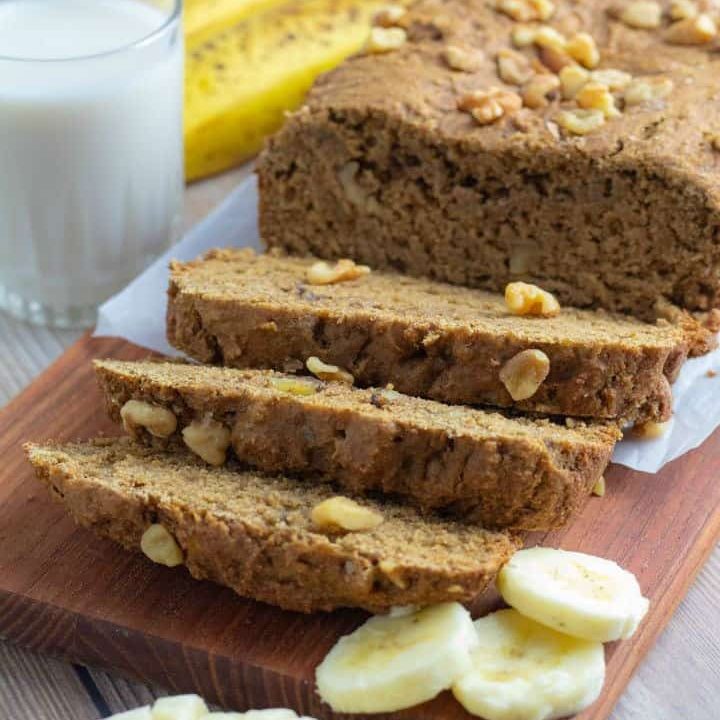 loaf of vegan banana bread sliced on cutting board with slices of banana and glass of milk