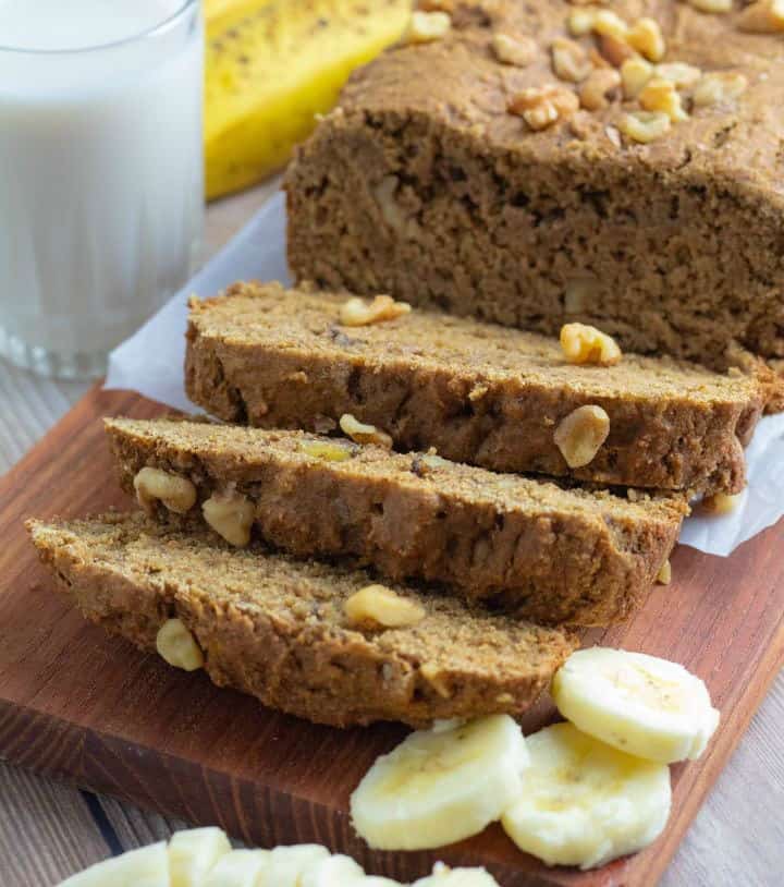 loaf of vegan banana bread sliced on cutting board with slices of banana and glass of milk