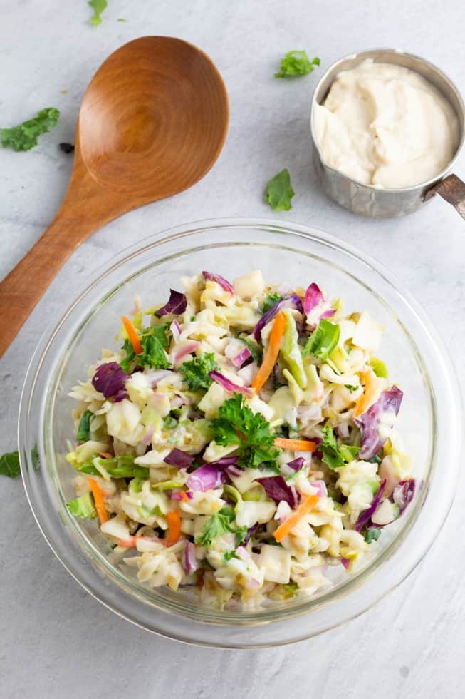 overhead shot of vegan coleslaw in glass bowl with wooden spoon and measuring cup with vegan mayo