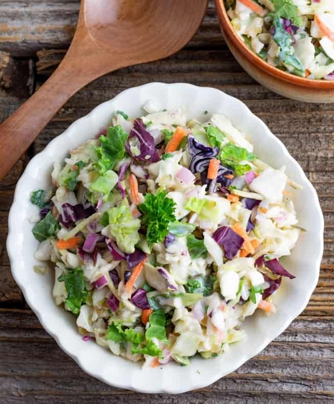 white scalloped bowl filled with vegan coleslaw on rustic wooden table with wooden spoon