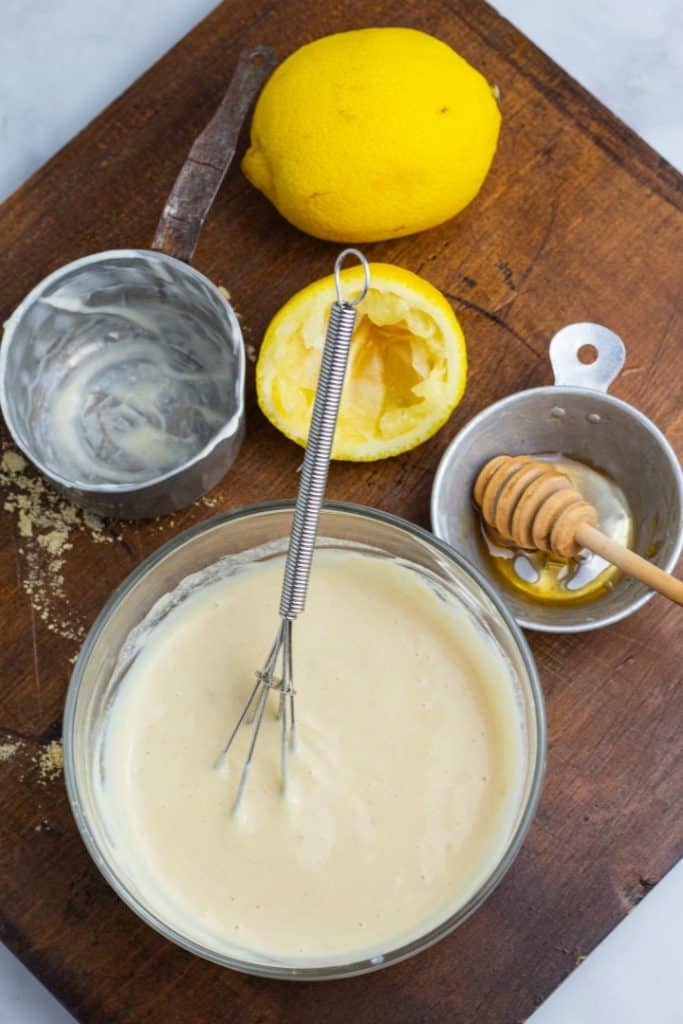 clear bowl of honey mustard sauce with whisk on wood board with squeezed lemon half and empty measuring cups