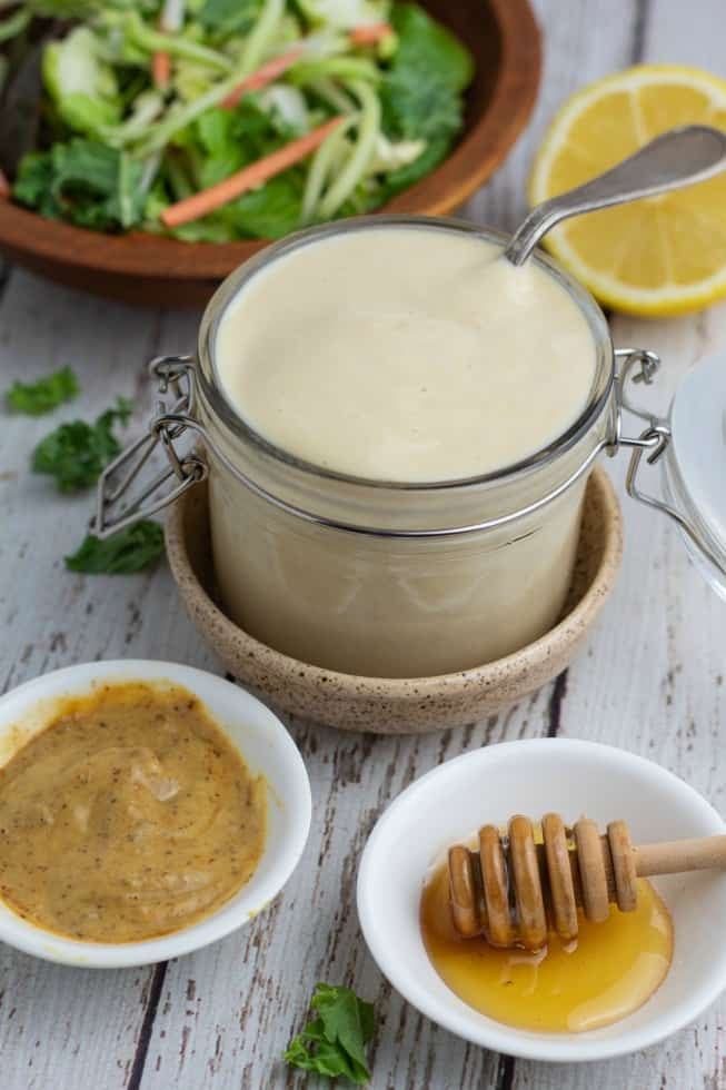 jar filled with honey mustard dressing and bent spoon with small white bowls of dijon mustard and honey setting in front