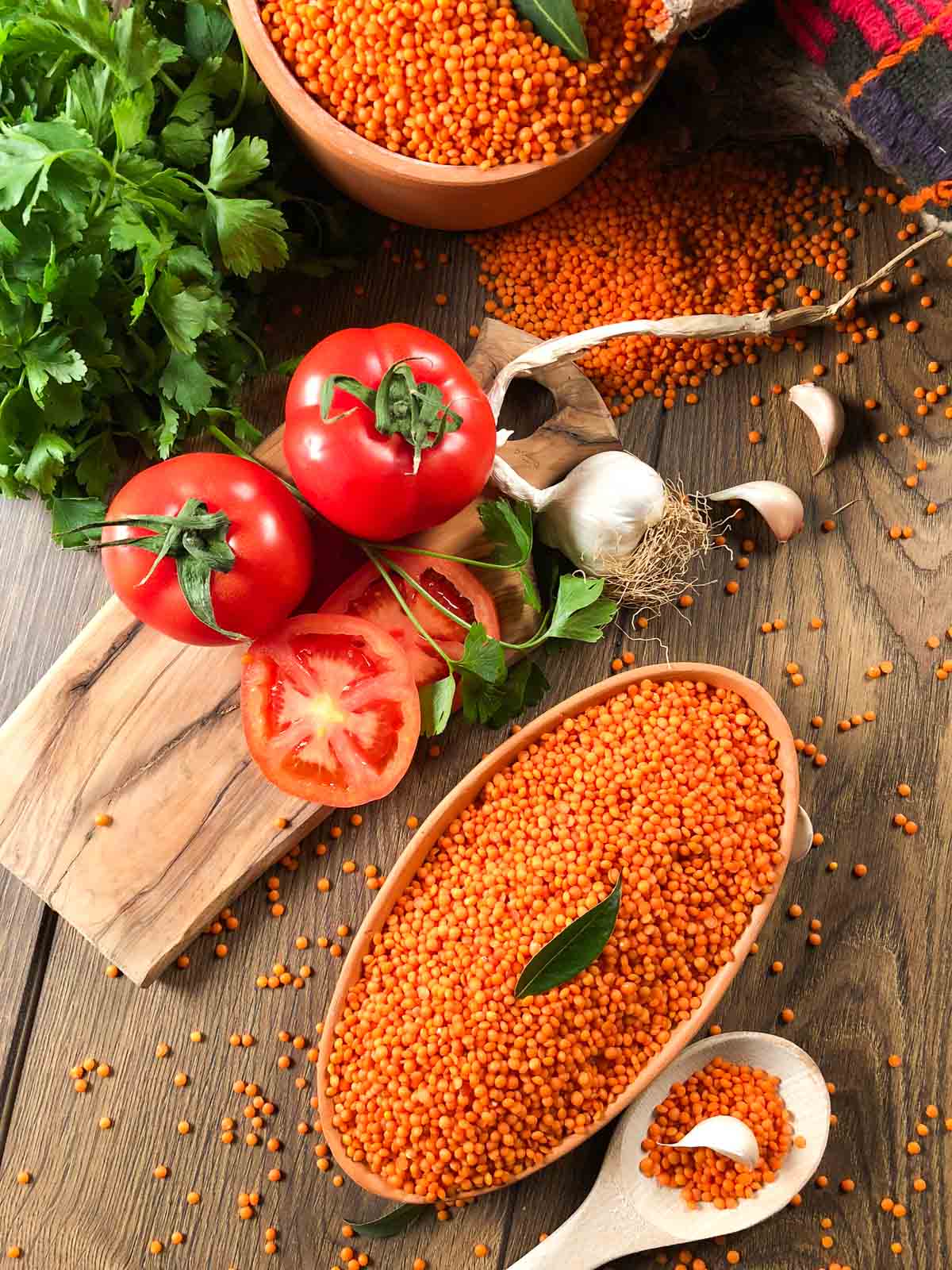 dry red lentils in oval wooden bowl with tomoatoes and garlic ingredients spread around.