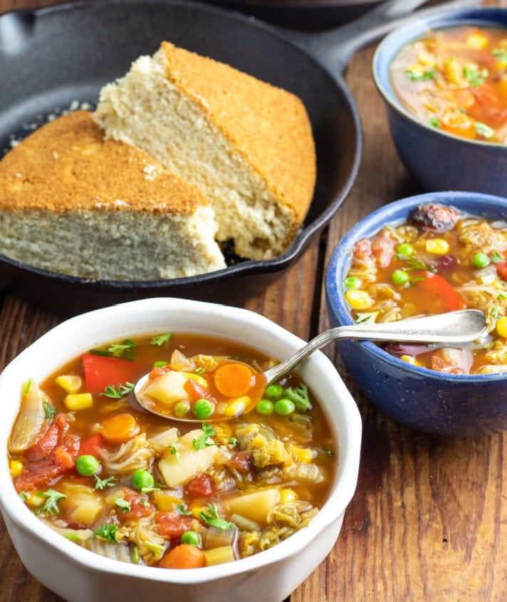 white bowl full of vegetable soup with spoon and cornbread in cast iron skillet