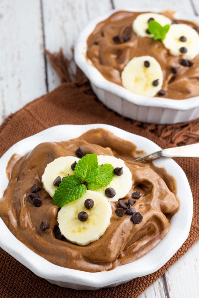 two white bowls with chocolate banana nice cream topped with sliced bananas, dairy free chocolate chips, and mint leaves