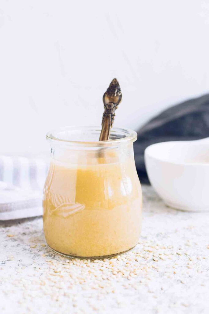 clear glass jar filled with homemade tahini sauce with a spoon inside jar.