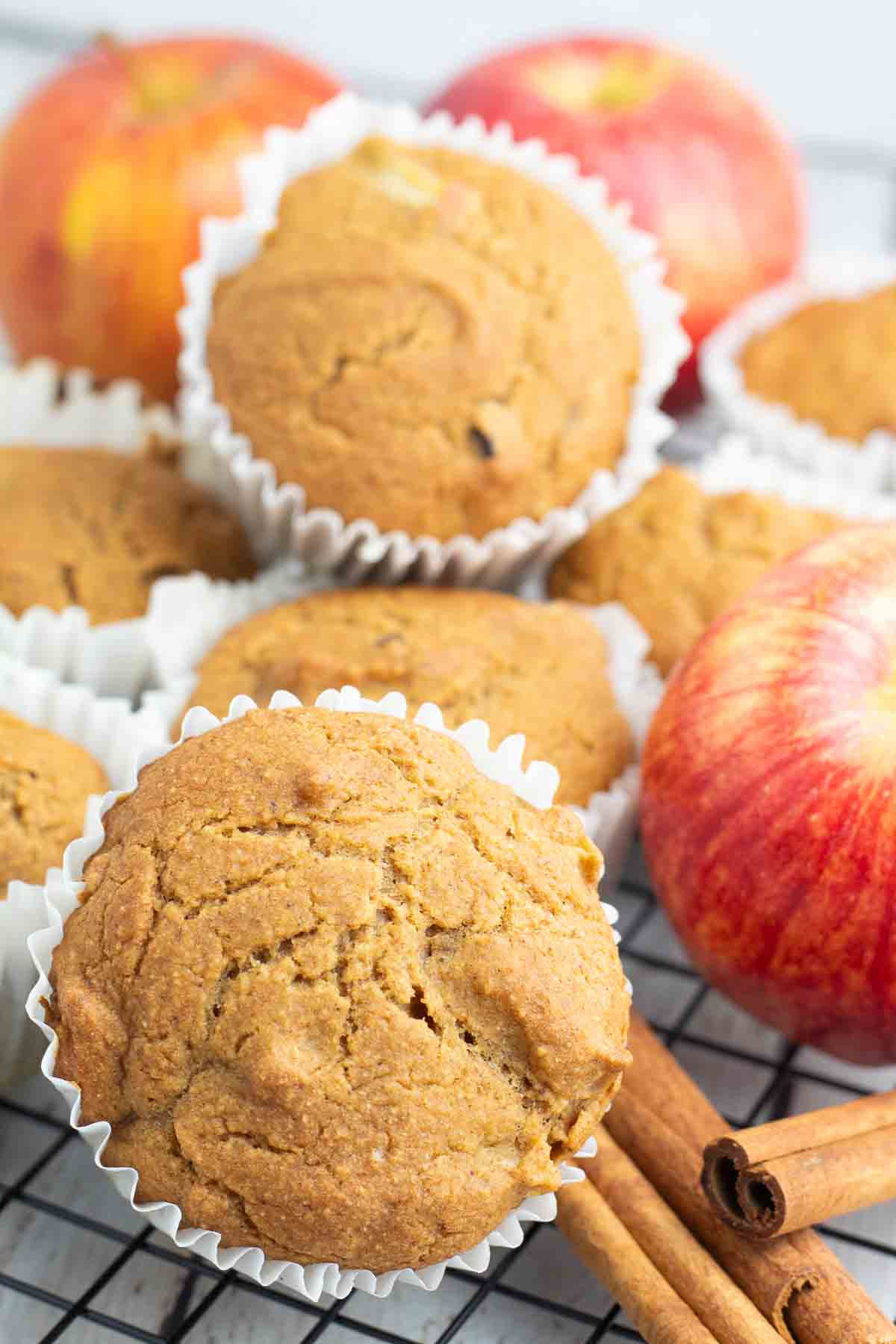 vegan applesauce muffins in white muffin cup paper with cinnamon sticks and apples surrounding them.