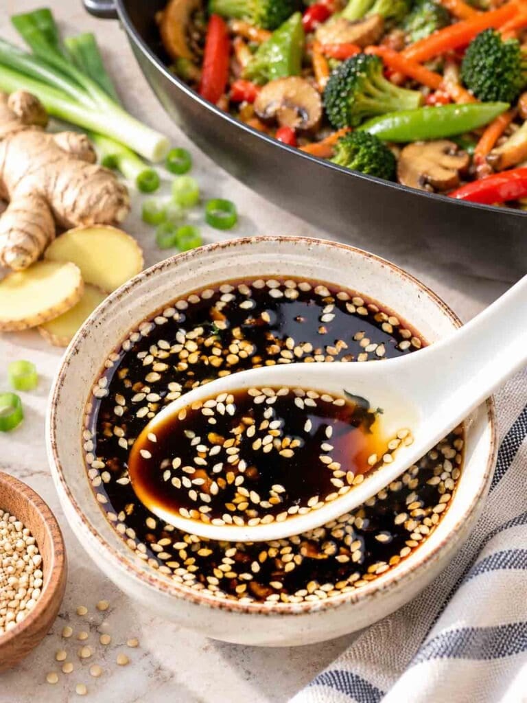 sweet ginger sesame sauce in a bowl with a spoon and a pan of stir fry veggies in background.