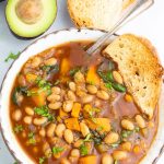 close up of bowl of white bean chili with avocado and crusty bread