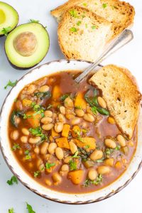 close up of bowl of white bean chili with avocado and crusty bread