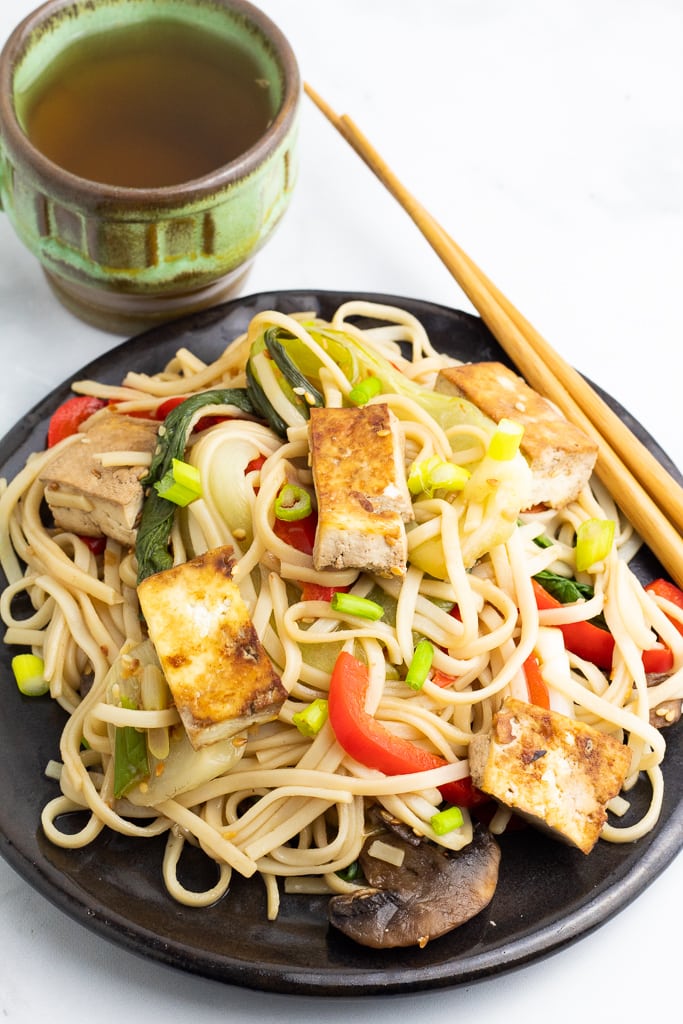 black plate filled with vegetable stir fry with tofu and chopsticks and hot tea
