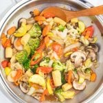 overhead shot stainless wok with stir fry veggies and wooden spoon on white background