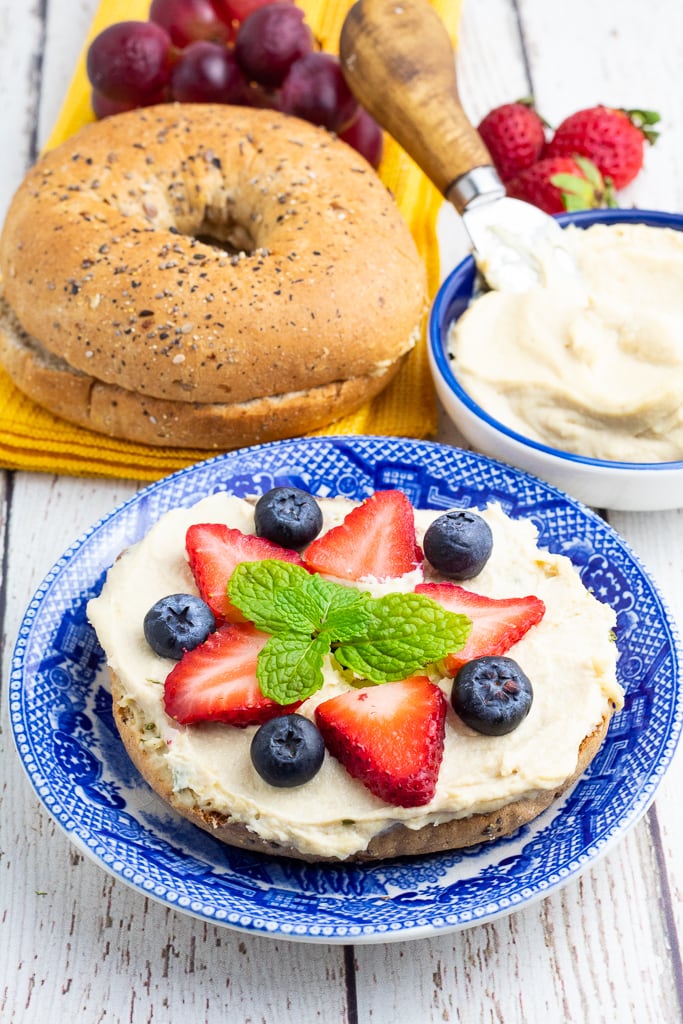 bagel half on bright blue plate covered with vegan cream cheese with strawberries, blueberries, and mint leaves