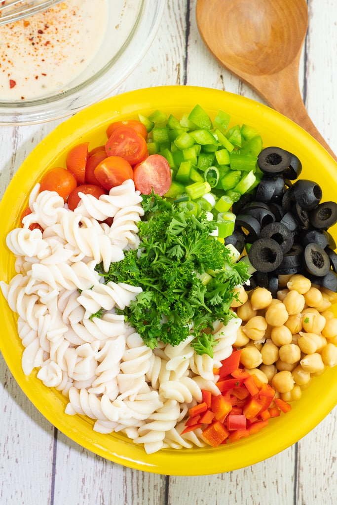 large yellow bowl with pasta salad ingredients, black olives, chickpeas, spiral pasta, and veggies