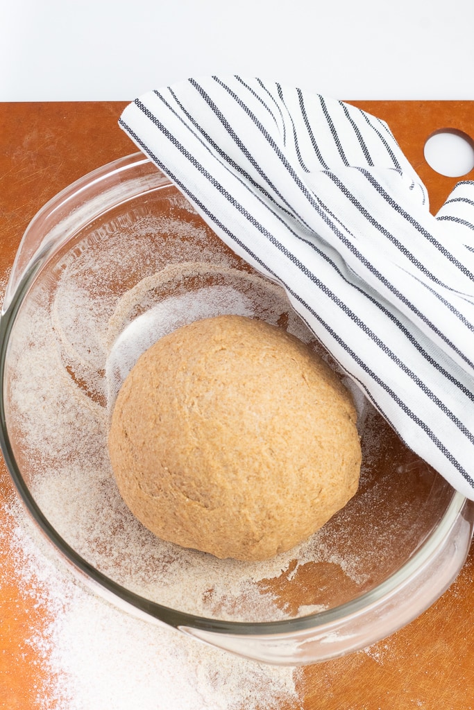 ball of bagel dough rising in glass bowl covered with dish towel