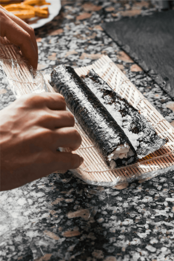 persons hands rolling sushi in nori with a sushi maker mat