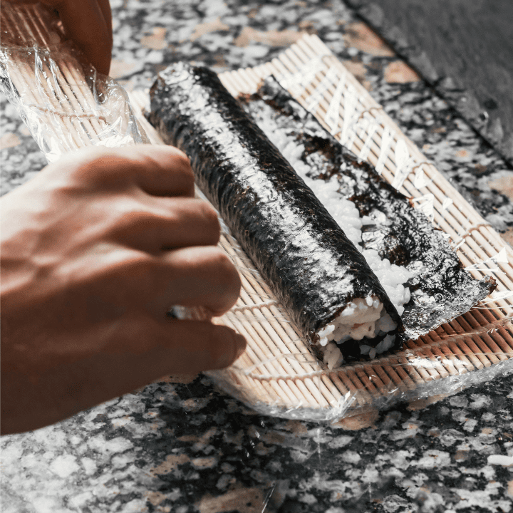persons hands rolling sushi in nori with a sushi maker mat