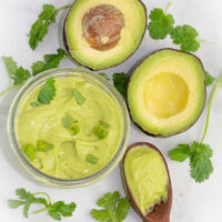 overhead shot of cilantro lime dressing in clear round bowl beside a sliced avocado and wooden spoon with the dressing on white background