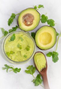 overhead shot of cilantro lime dressing in clear round bowl beside a sliced avocado and wooden spoon with the dressing on white background