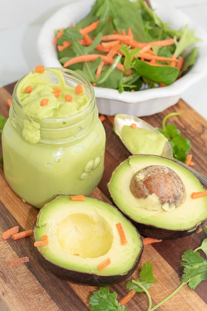 small jar of avocado cilantro lime dressing with a sliced avocado in front and salad in background