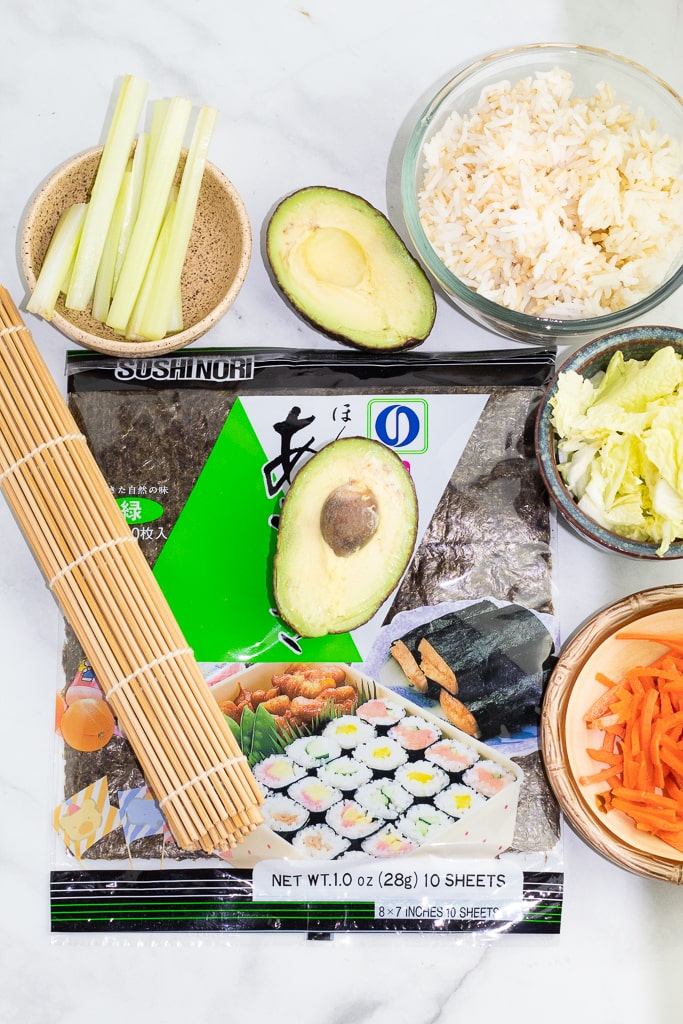 overhead shot of nori sheets, sliced carrots, celery, avocado, bowl of sushi rice and bamboo mat