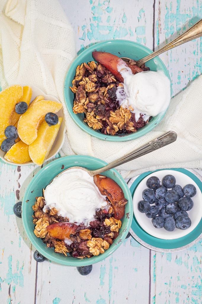 overhead photo of 2 blue bowls filled with blueberry crisp and vanilla ice cream with bowl of blueberry and peaches on sides