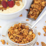 small glass decorative jar filled with gluten free granola and spoon. background scoop of granola and yogurt bowl