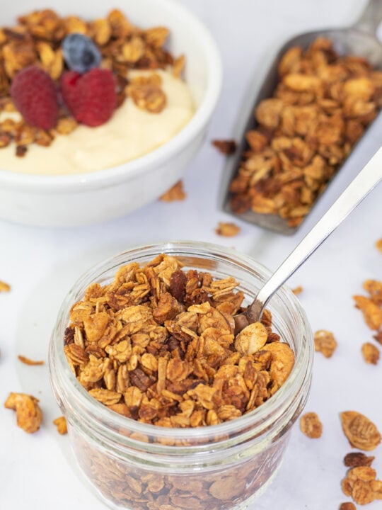 small glass decorative jar filled with gluten free granola and spoon. background scoop of granola and yogurt bowl