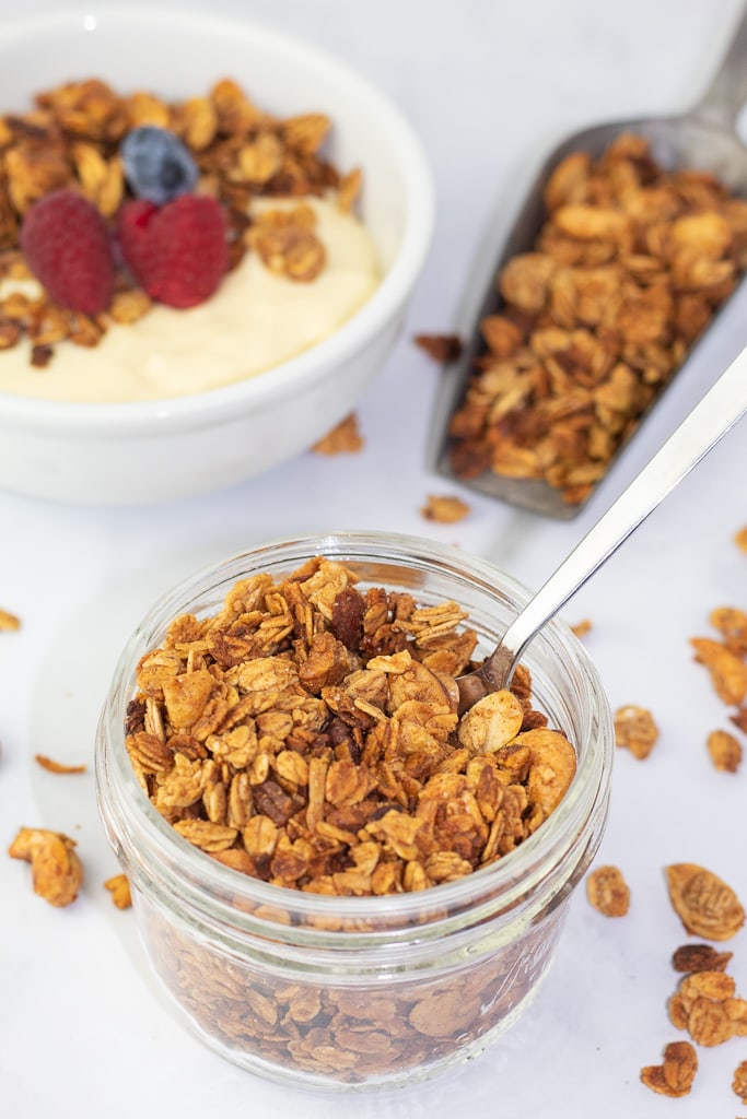 small glass decorative jar filled with gluten free granola and spoon. background scoop of granola and yogurt bowl