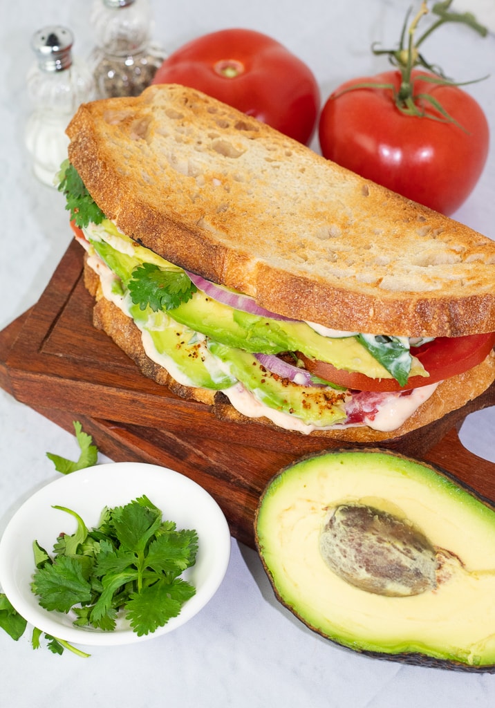 avocado sandwich on stacked wooden boards surrounded by cilantro, tomatoes, avocados and salt and pepper
