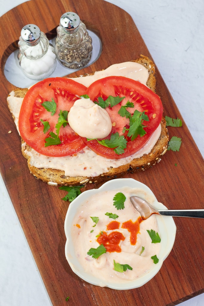 white bowl of spicy vegan mayo with hot sauce and a tomato sandwich and salt and pepper shakers