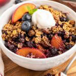 blueberry peach crisp in a white bowl with a scoop of vanilla nice cream.